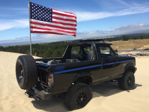 1992 Ford Bronco - lifted black Ford Bronco flying an American flag sits on sand dunes, spare tire mounted at the rear.