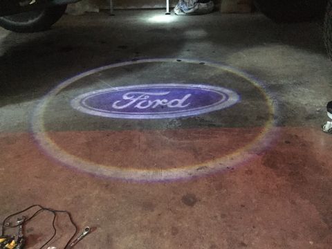 1992 Ford Bronco - A Ford logo is projected onto a concrete floor, likely from a vehicle door light. Tools and wiring are visible in the lower left corner.