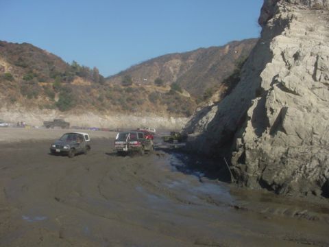 1988 Ford Bronco - Several off-road vehicles are stuck in thick mud near a rocky embankment under a clear blue sky.