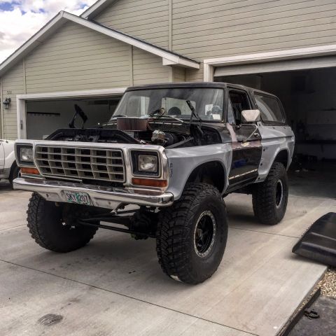 1979 Ford Bronco - Late-1970s Ford Bronco in primer black-and-silver, hood off exposing the engine, lifted on black wheels and mud tires, parked in a resident…