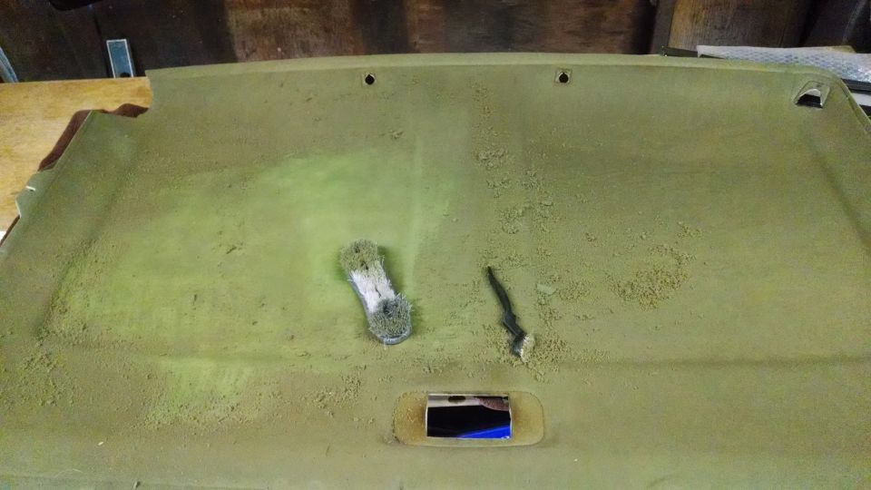 Removed green headliner panel covered in dust being cleaned with a wire brush and scrub pad in a workshop