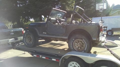 1985 Jeep CJ7 - A dark colored Jeep CJ7 is loaded onto a flatbed trailer, hitched to a pickup truck. The Jeep is open-topped with a roll cage visible.