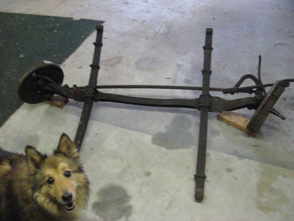 Rusty front axle assembly with brake drum and suspension components, with a dog in the foreground.