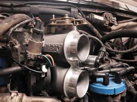 1995 Ford Bronco - A close-up view of a modified engine bay, focusing on a silver BBK throttle body with dual inlets. Various hoses, wires, and a blue distrib…