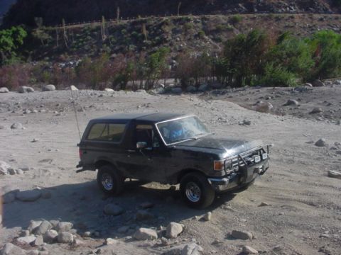1988 Ford Bronco - A black Ford Bronco is parked on a dirt road with rocky terrain and sparse vegetation in the background.