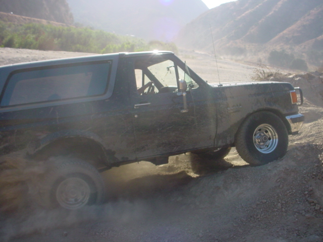 Mud-covered dark blue Ford Bronco climbing a dusty off-road hill, seen from the passenger side
