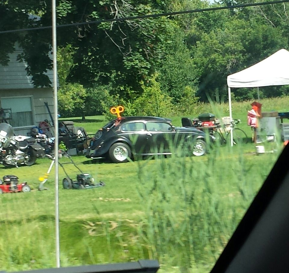 Black Volkswagen Beetle with a wind-up toy key on its roof, parked on a grassy lawn next to a motorcycle and lawnmowers