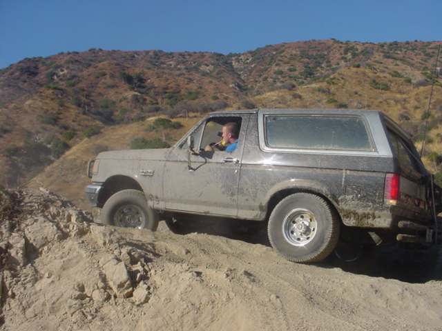 Muddy Ford Bronco climbing a rocky dirt trail in arid hills, driver visible in window