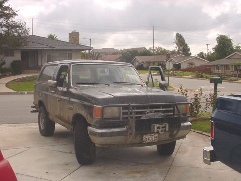 1988 Ford Bronco - A dirty black Ford Bronco is parked in a suburban driveway, with its driver's door open.