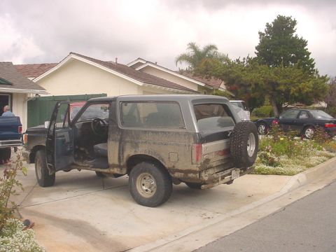 1988 Ford Bronco - A muddy 1988 Ford Bronco sits in a driveway with its doors open and a spare tire on the back.