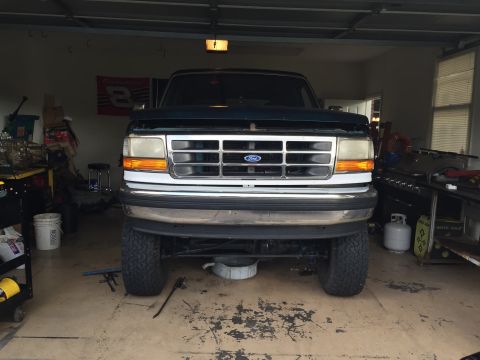 1994 Ford Bronco - The front view of a dark green Ford Bronco is parked inside a garage. The vehicle has a chrome grille and bumper, and large off-road tires…