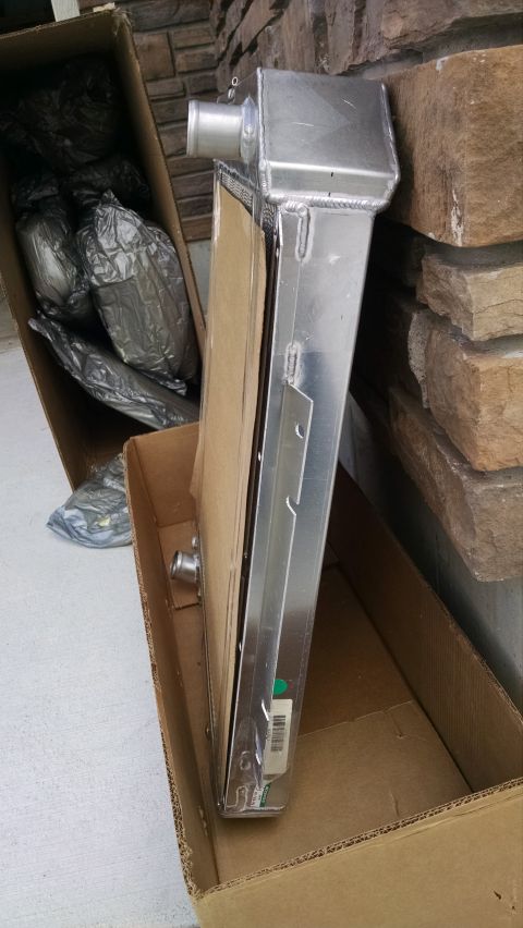 1979 Ford F-150 - A new aluminum radiator is shown partially unpacked from its cardboard box, resting against a stone wall. The radiator has silver inlet and…
