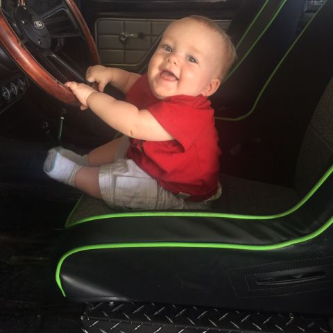 1979 Ford Bronco - A smiling baby sits in the driver's seat of a vehicle, holding onto the steering wheel. The baby is wearing a red shirt and light-colored s…