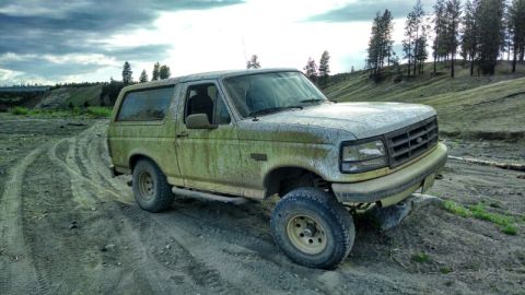 1992 Ford Bronco - A dirty, light-colored 1992 Ford Bronco is parked on a dirt road with tire tracks. The vehicle is positioned at a 3/4 front angle, facing t…