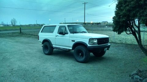 1992 Ford Bronco - A white two-door Ford Bronco is parked on a gravel lot under a cloudy sky. The SUV has large off-road tires and appears to be lifted.