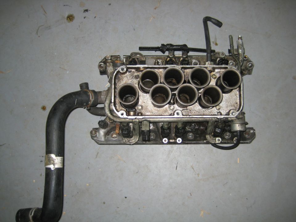 Engine intake manifold with eight runners and a black hose attached, on a concrete floor.