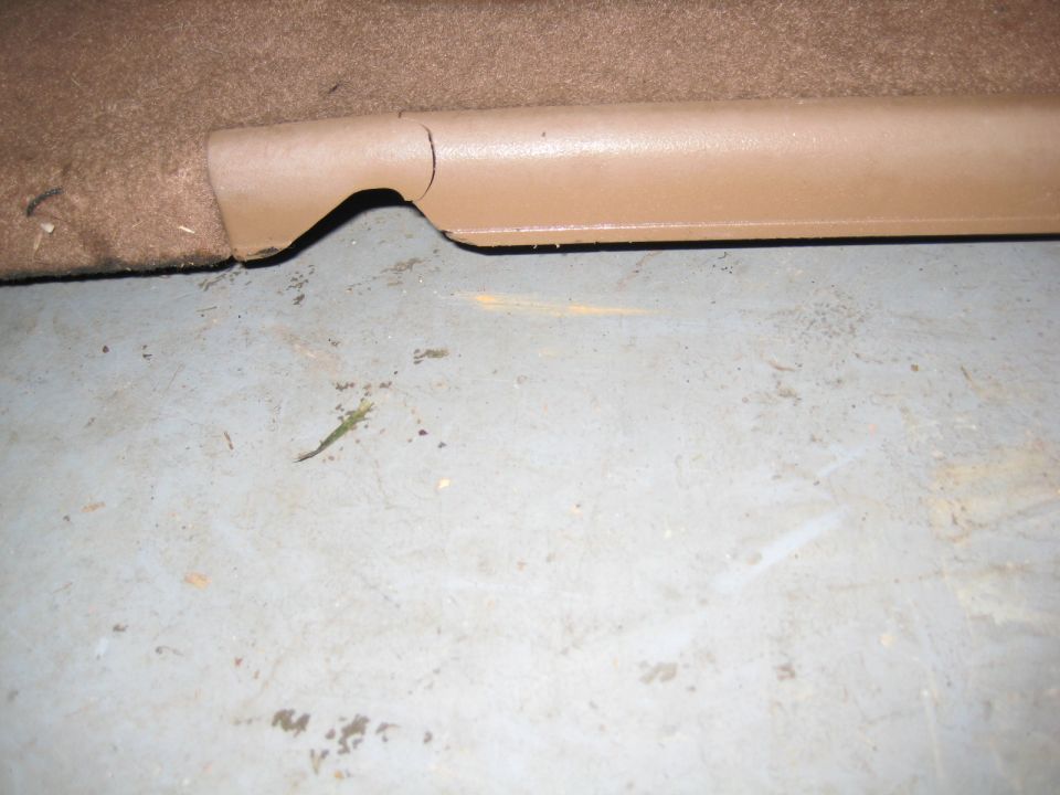 Tan plastic trim piece with crack next to brown carpet and grey floor.