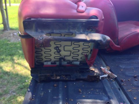 1979 Ford Bronco - A red car seat with exposed springs and a rusted metal mounting bracket is shown, resting on the dark bed of a pickup truck.