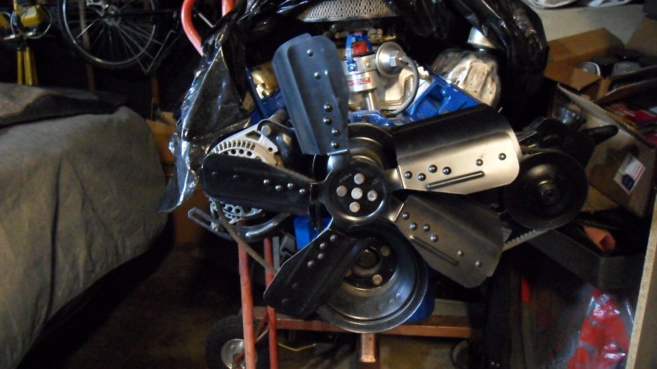Black metal fan with four blades on a blue engine with an alternator.