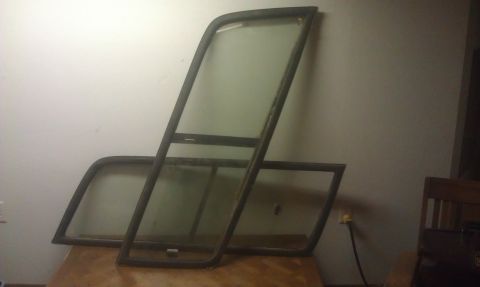 1992 Ford Bronco - Two framed glass windows, likely for a vehicle, are leaning against a wall on a wooden surface. One window is taller and more upright, whil…