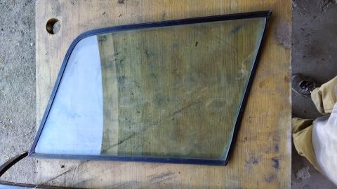 1992 Ford Bronco - A triangular automotive side window with a black rubber seal is laid on a wooden surface next to a concrete floor. A person's legs wearing…