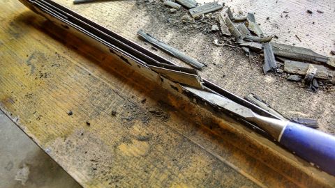 1992 Ford Bronco - A close-up shot shows a chisel with a blue handle being used to scrape away material from a metal object on a wooden surface. Debris, likel…