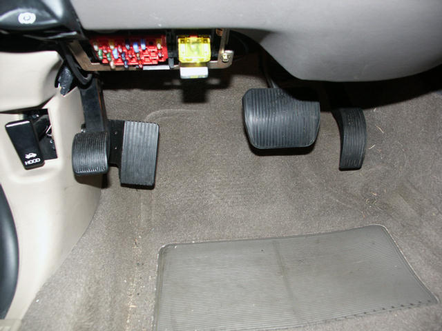 Car footwell with brake and gas pedals, exposed fuse box and hood release above.