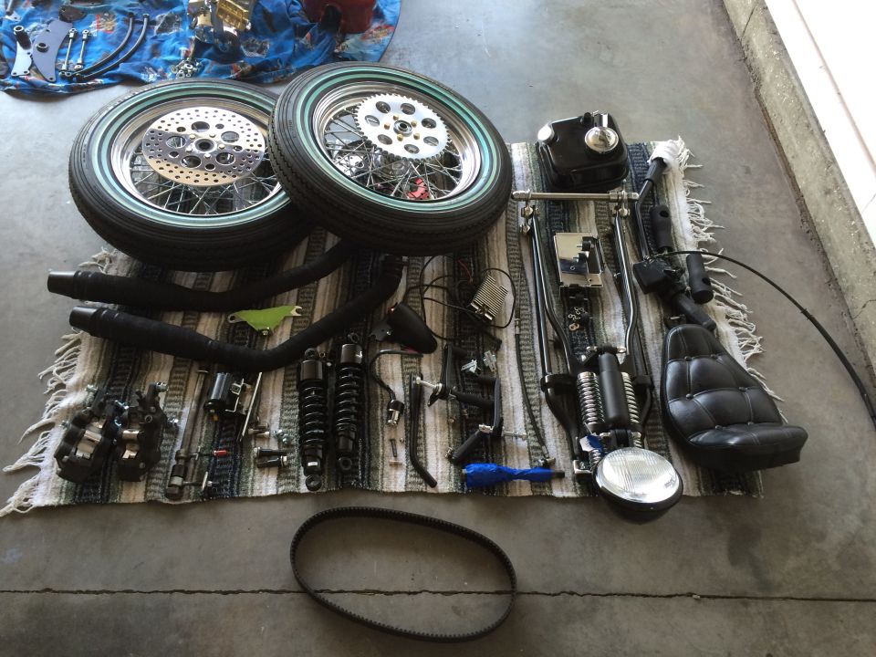 Motorcycle parts including wheels, exhaust, seat, shocks, and headlight laid out on a rug.