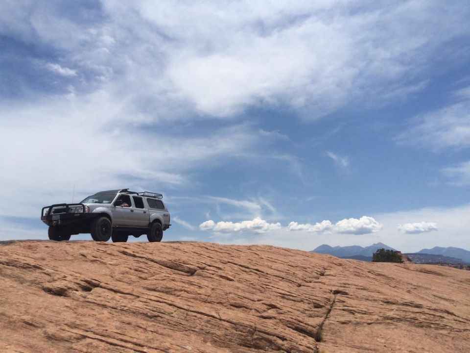 Silver Nissan Frontier pickup truck with bull bar and roof rack on rocky desert terrain.