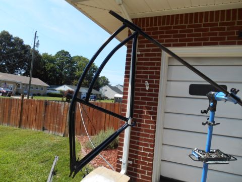 Black bicycle frame on a blue repair stand in front of a brick garage.