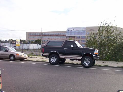 1996 Ford Bronco - A black Ford Bronco is parked on the side of the road next to a tan sedan, with a large building in the background.