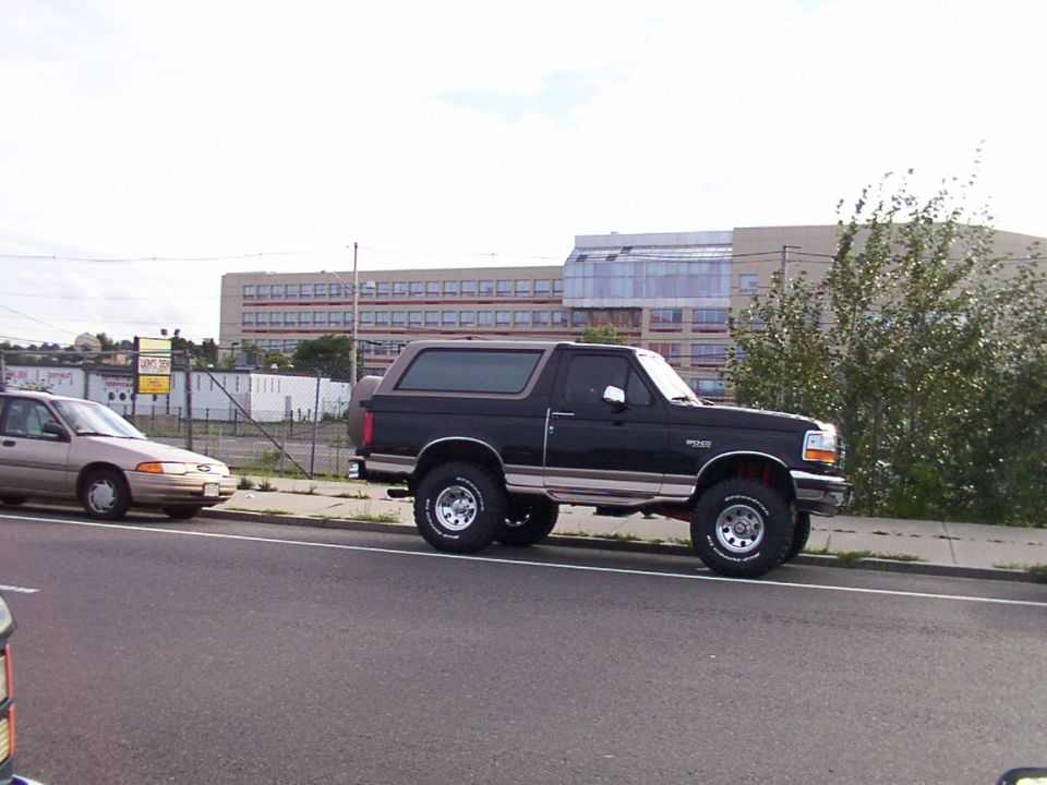 Black Ford Bronco parked on street, tan sedan next to it. Building in background