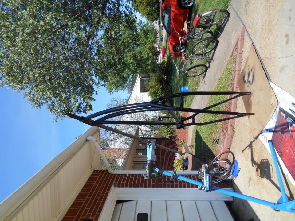 Black bicycle frame on a blue repair stand in a driveway.
