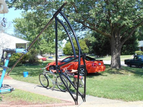 Orange Ford Mustang parked in driveway with bicycle repair stand and green pickup truck.
