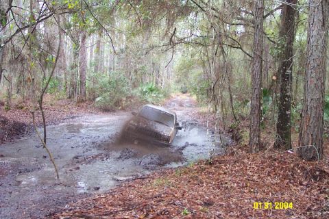 1985 Ford Bronco - Off-road vehicle struggling through a deep mud puddle on a forest trail.