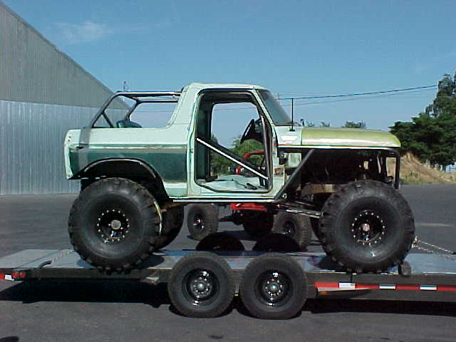Stripped pale green Bronco trail rig with roll cage and big lug tires strapped to a flatbed trailer