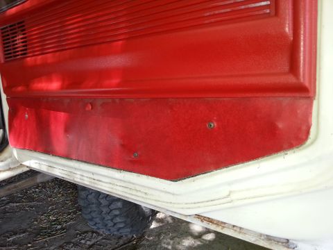 1978 Ford Bronco - Close-up view of the red interior door panel of a vehicle, showing some wear and tear. A portion of a black tire is visible at the bottom.