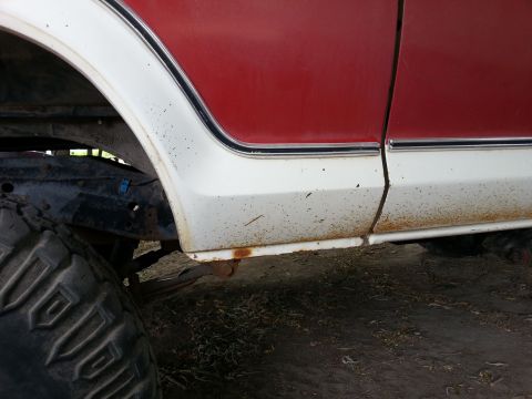 1978 Ford Bronco - A close-up, side view of the lower body of a red and white vehicle, showing rust and dirt along the bottom edge and wheel well.
