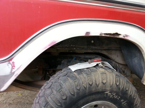 1978 Ford Bronco - Close-up view of the rear wheel well of a red vehicle, showing significant rust and damage to the fender flare. A screwdriver rests on a pi…