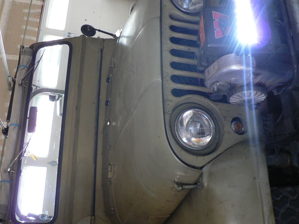 Vintage Jeep M38A1 driver's side front fender, grille, and door with bright light shining from engine bay.