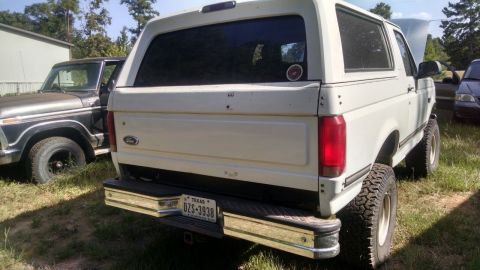 1996 Ford Bronco - A white Ford Bronco is parked next to another vehicle, showing its rear end and lifted suspension.