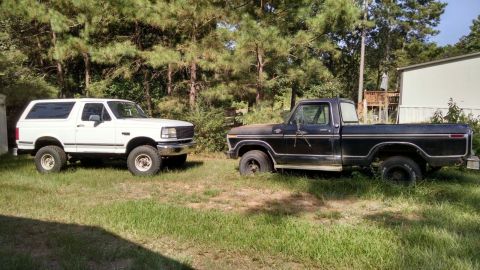 1996 Ford Bronco - Two Ford vehicles, a Bronco and a pickup truck, sit parked in a yard with trees and a building in the background.
