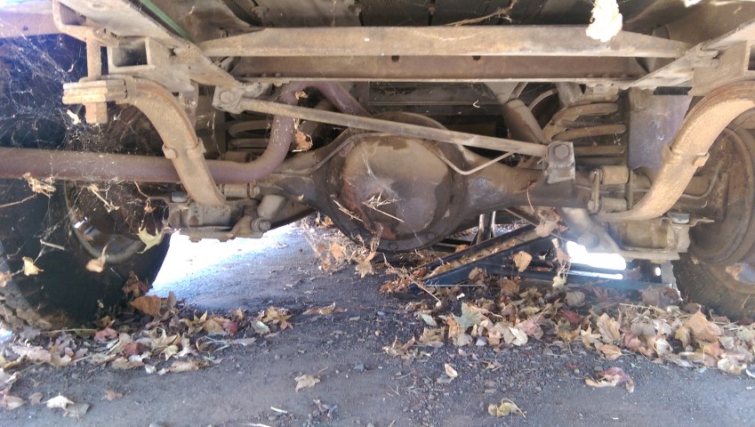 Underside of a 1967 Chevrolet C/K Pickup truck showing the rear axle, leaf springs, and exhaust pipes, covered in cobwebs and fallen leaves
