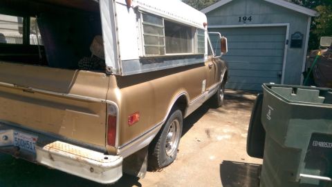 1968 Chevrolet C/K Pickup - A gold 1968 Chevrolet C/K Pickup truck with a camper shell is parked in front of a garage.