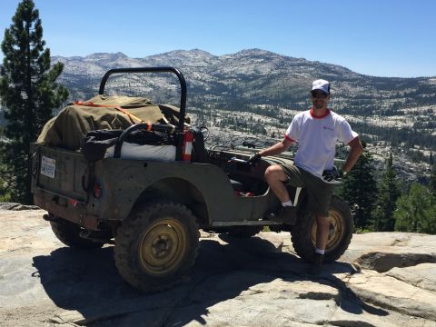 1946 Jeep CJ5 - A man stands next to a vintage Willys Jeep parked on a rocky terrain with mountains in the background.