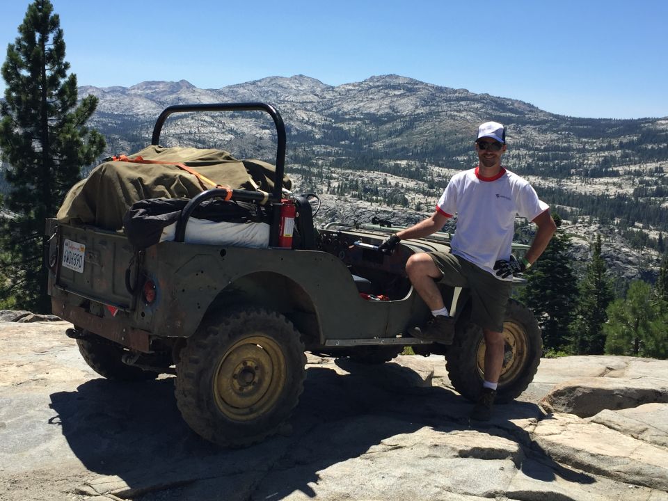Man posing with a green Willys CJ-3A loaded with gear on a rocky mountain overlook