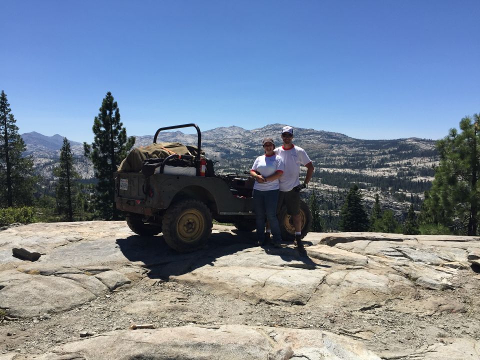 Olive green Jeep with spare tire on the back, parked on a rocky mountain landscape with two people beside it