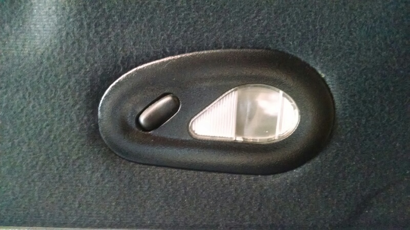 Black oval interior dome light with a push button and a clear ribbed lens, mounted on dark grey fabric