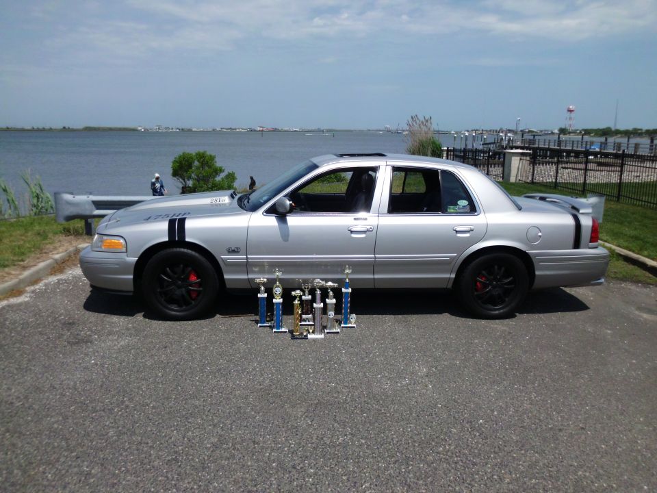 Silver sedan with black racing stripes on hood and trunk, black wheels, red brake calipers, and a rear spoiler. Trophies are displayed in front of the car