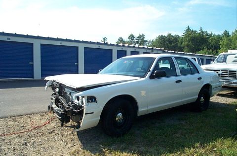 2001 Ford Crown Victoria - A white sedan, possibly a 2001 Ford Crown Victoria, sits damaged in front of a row of blue storage unit doors.